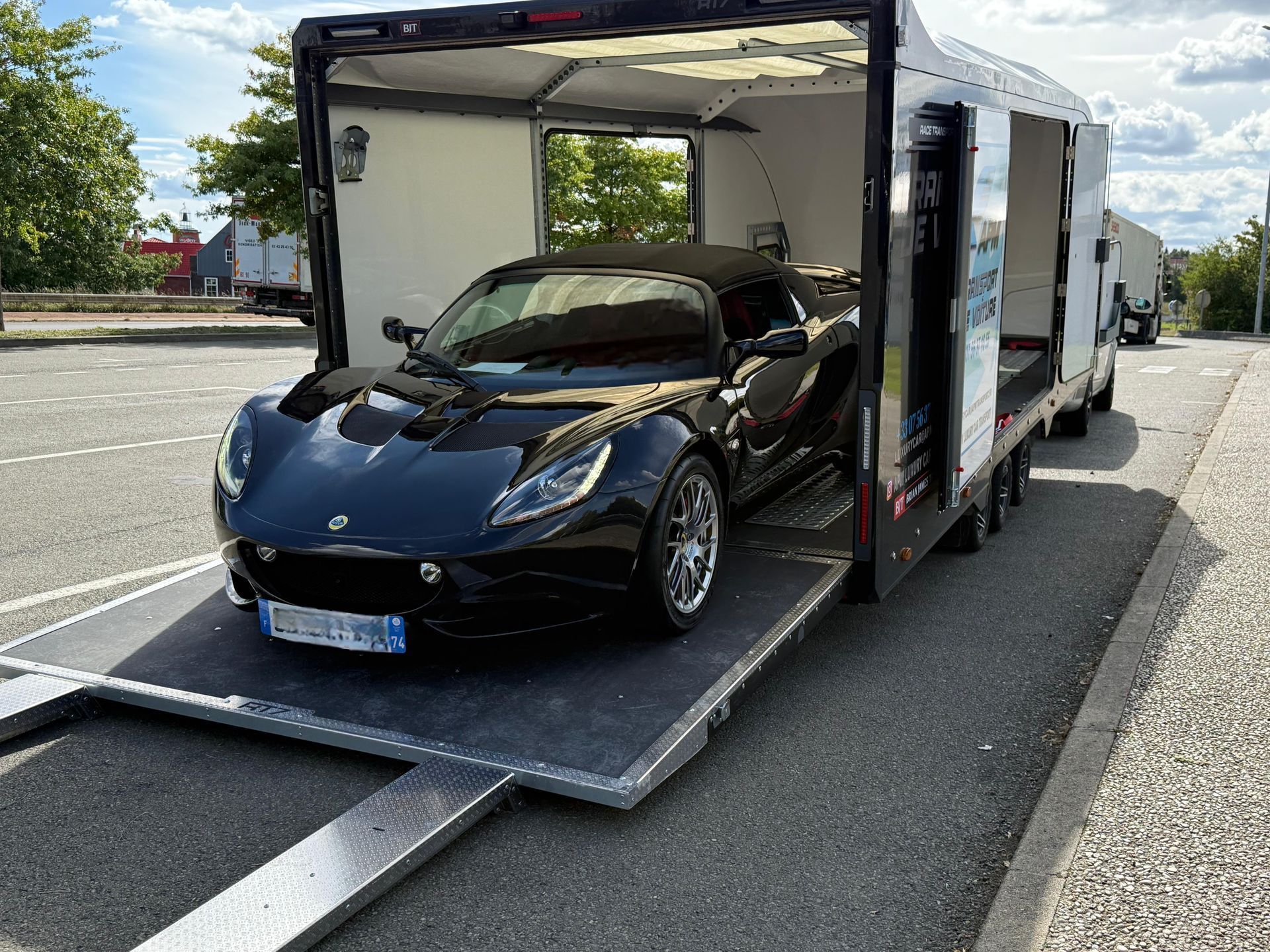 Une Lotus noire, voiture de sport, est garée sur la rampe abaissée d'une remorque porte-voitures, sur un parking.