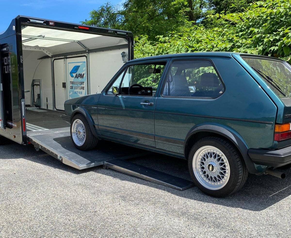 Une Volkswagen Golf vintage turquoise garée sur la rampe métallique d'une remorque porte-voitures noire ouverte.