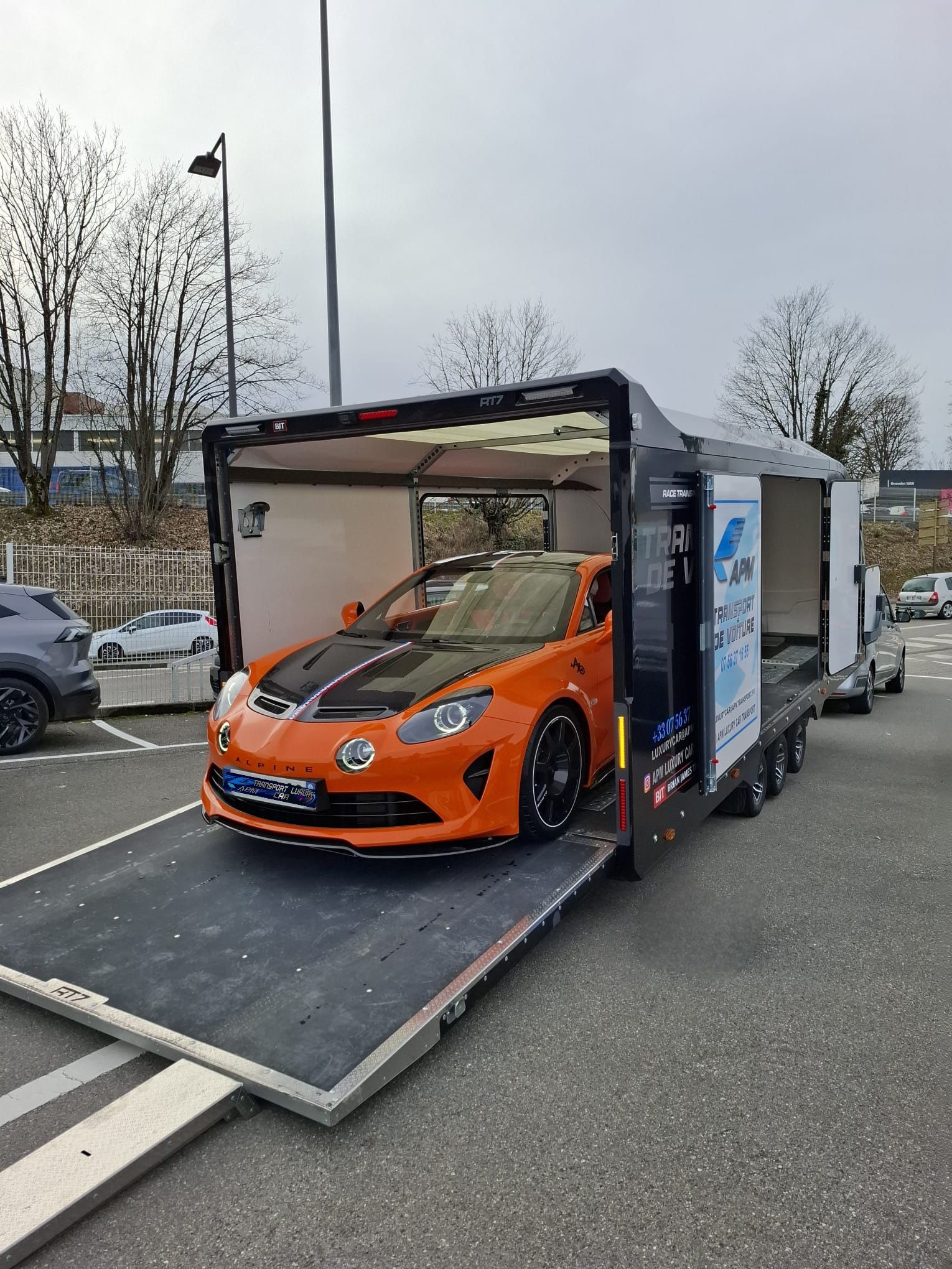 Une voiture de sport Alpine A110 orange est chargée dans une remorque porte-voitures noire fermée sur un parking.
