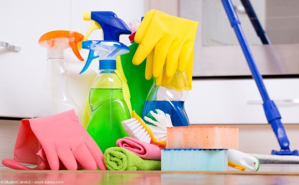 Bunte Reinigungsmittel auf einem Tisch, darunter Sprühflaschen, Handschuhe, Schwämme und Bürsten.