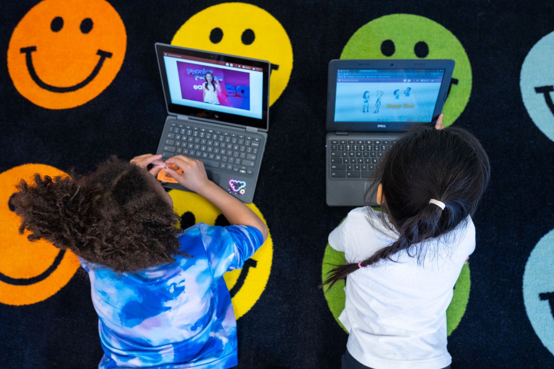 Two children on laptops on a smiley face rug. One girl has curly hair, the other a braid.