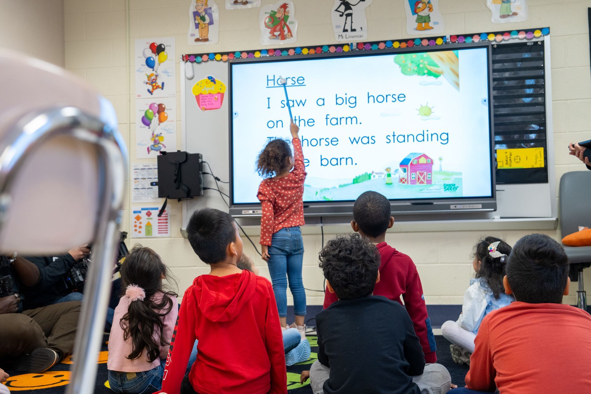 Children in a classroom, pointing at a smartboard displaying a sentence about a horse on a farm.