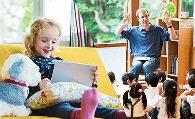 A little girl is sitting on a couch reading a book while a man is sitting in front of a group of children.