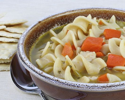 Un tazón de sopa de pollo con fideos con zanahorias y galletas de fondo.