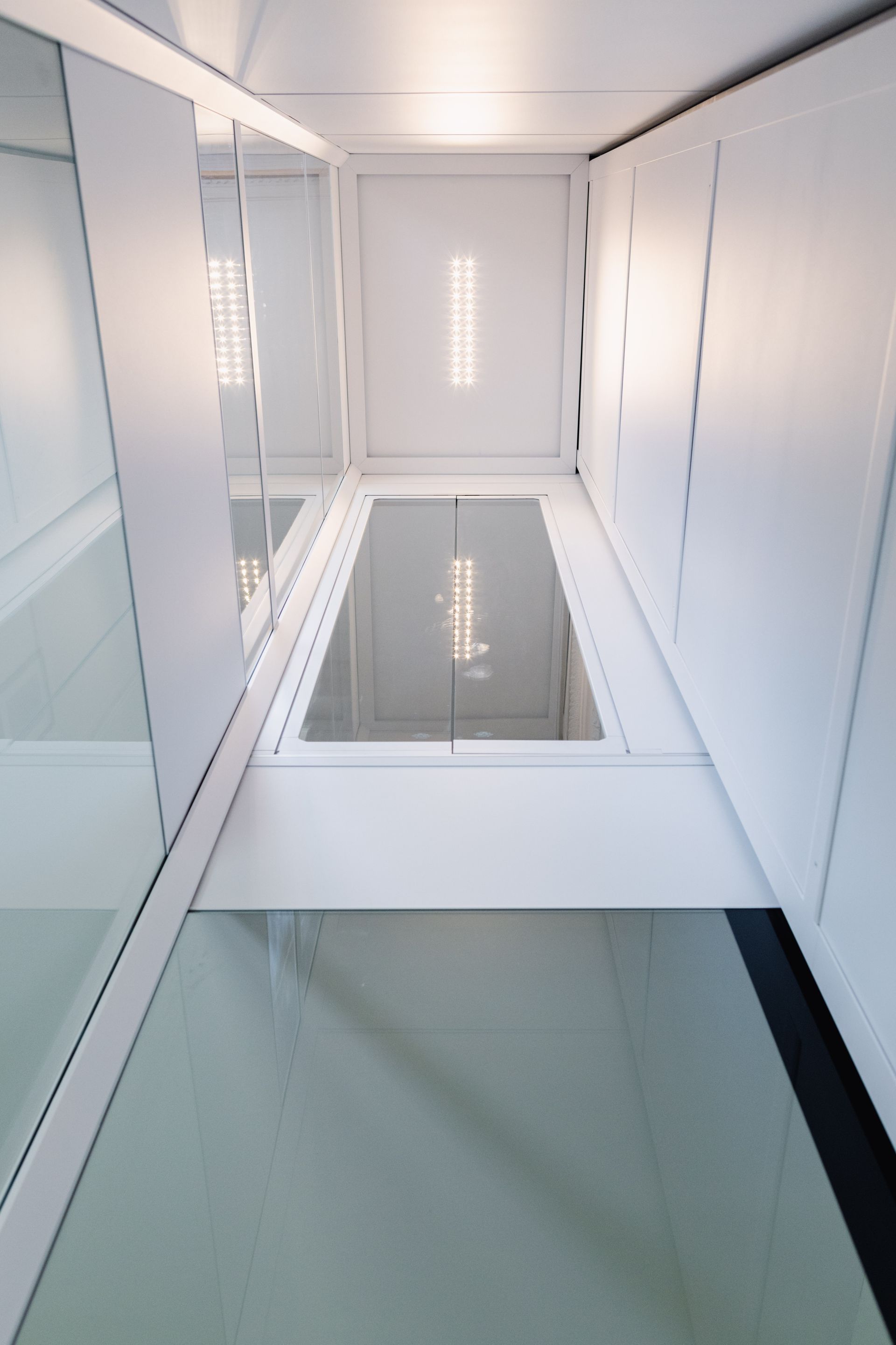 Le regard s'élève au-dessus d'un étroit couloir blanc. Sol et murs en verre, lumière vive au plafond.