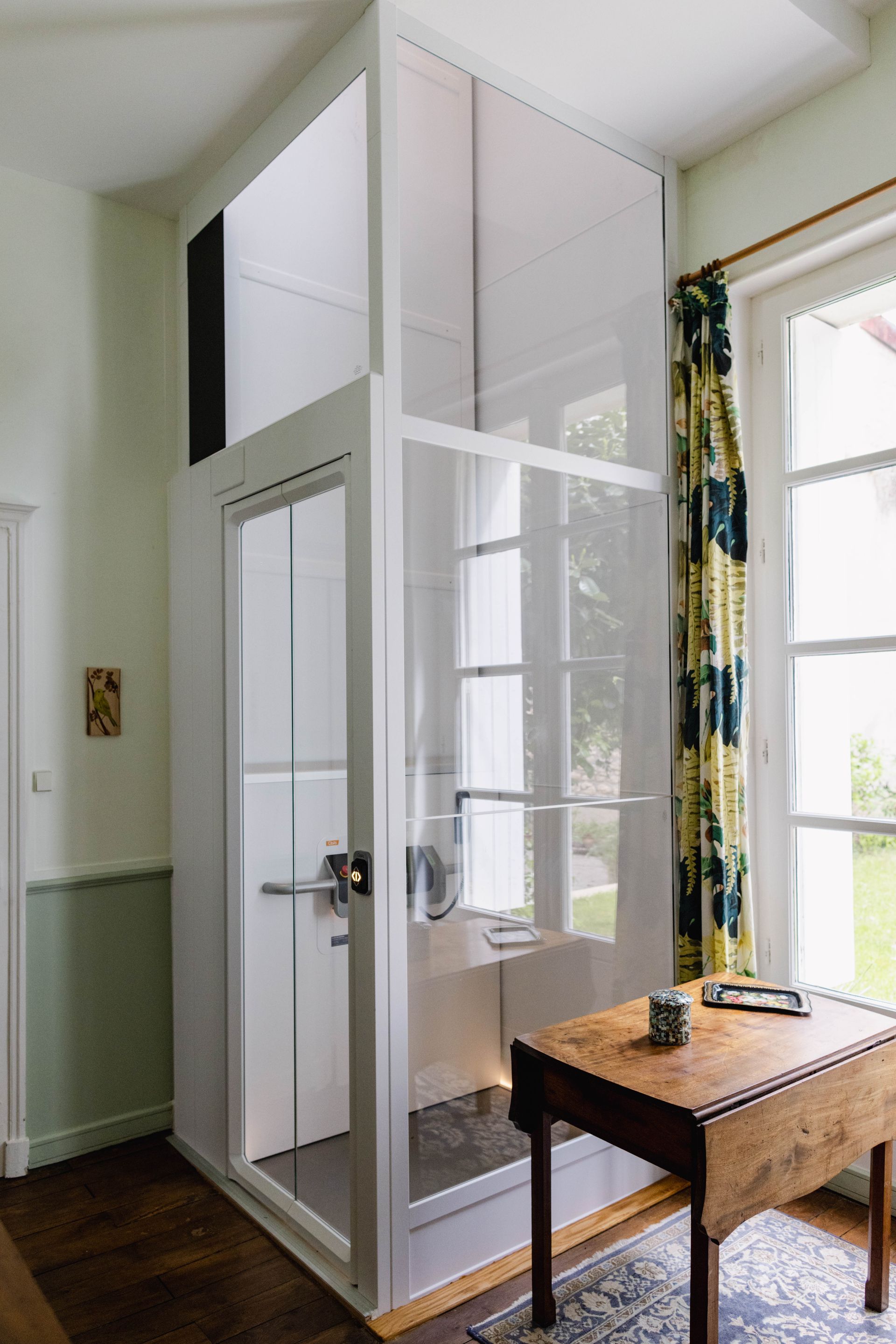 Ascenseur intérieur pour domicile avec panneaux de verre et blancs, à côté d'une fenêtre et d'une table.