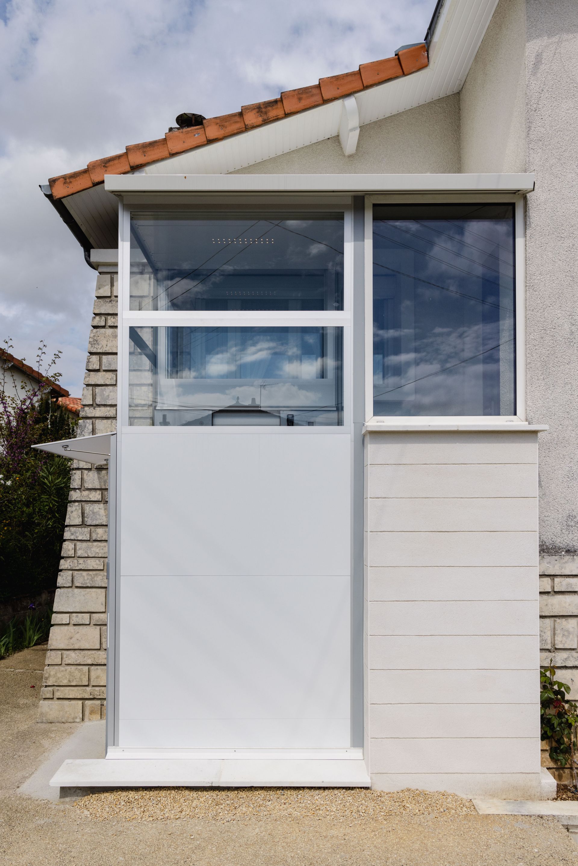 Porche d'entrée blanc avec fenêtres en verre, porte blanche et éléments en pierre sous un toit de tuiles.