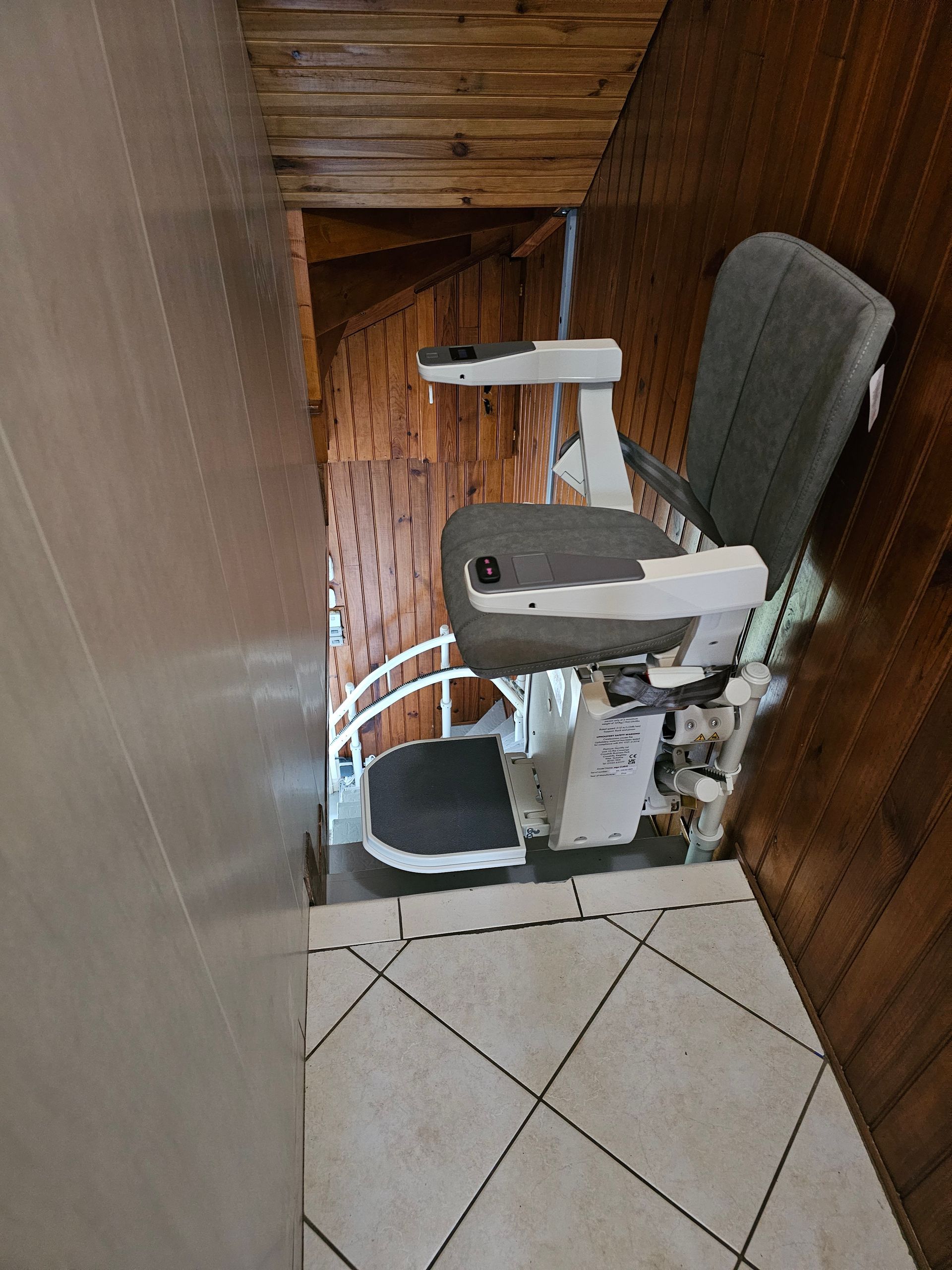 Monte-escalier sur un escalier en bois. L'ascenseur est gris et blanc. Sol carrelé en dessous.