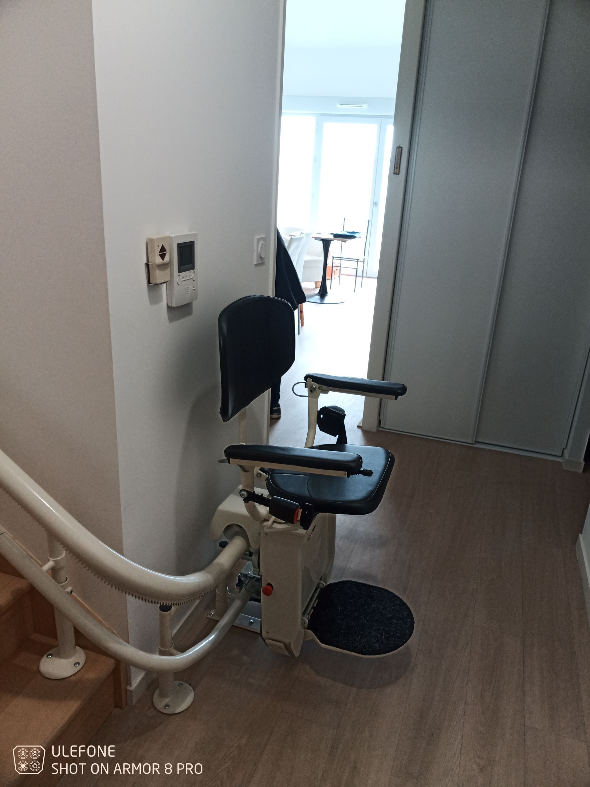 Un monte-escalier est installé sur un escalier adossé à un mur blanc. Le siège du monte-escalier est noir et blanc.