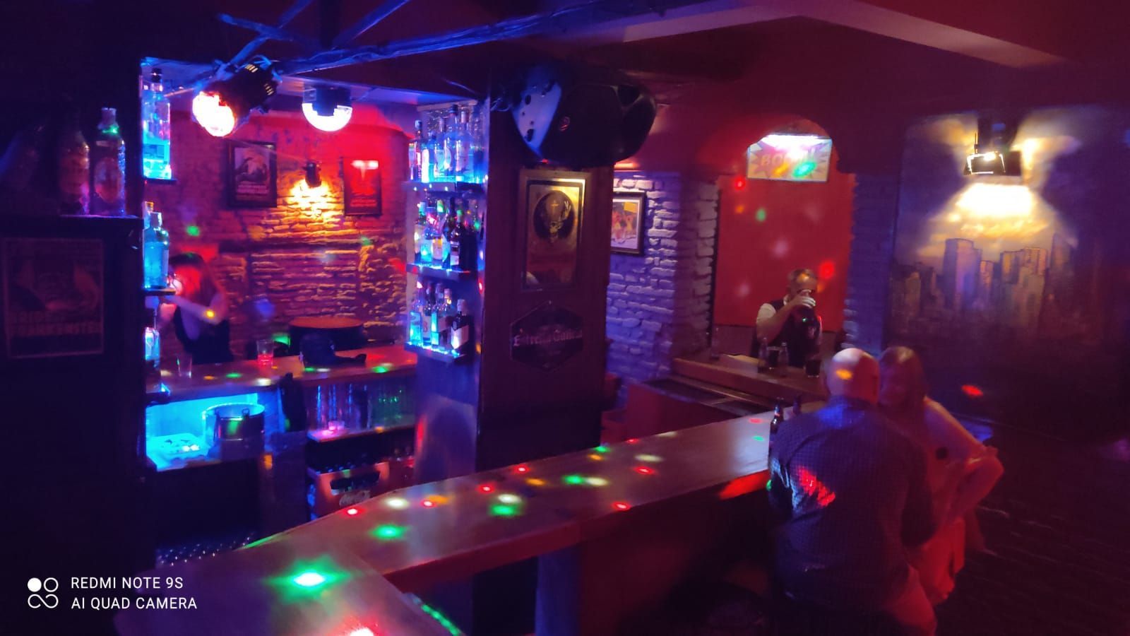 Vista interior de un bar con luces de colores, barra y gente.