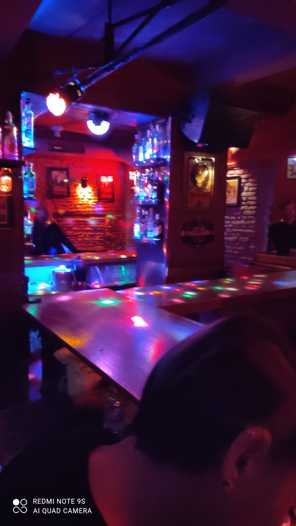 Un bar tenuemente iluminado con un camarero. Luces de colores se reflejan en el mostrador y las botellas.