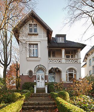 Ein großes weißes Haus mit Treppen, die zur Haustür hinaufführen, umgeben von Büschen und Bäumen.