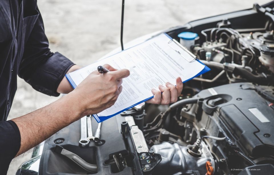 Ein Mechaniker macht sich Notizen auf einem Klemmbrett, während er in einer Werkstatt einen Automotor untersucht.