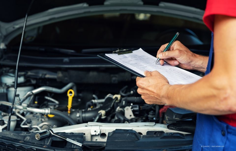 Ein Mechaniker in blauem Overall schreibt auf einem Klemmbrett, während er einen offenen Automotor untersucht.