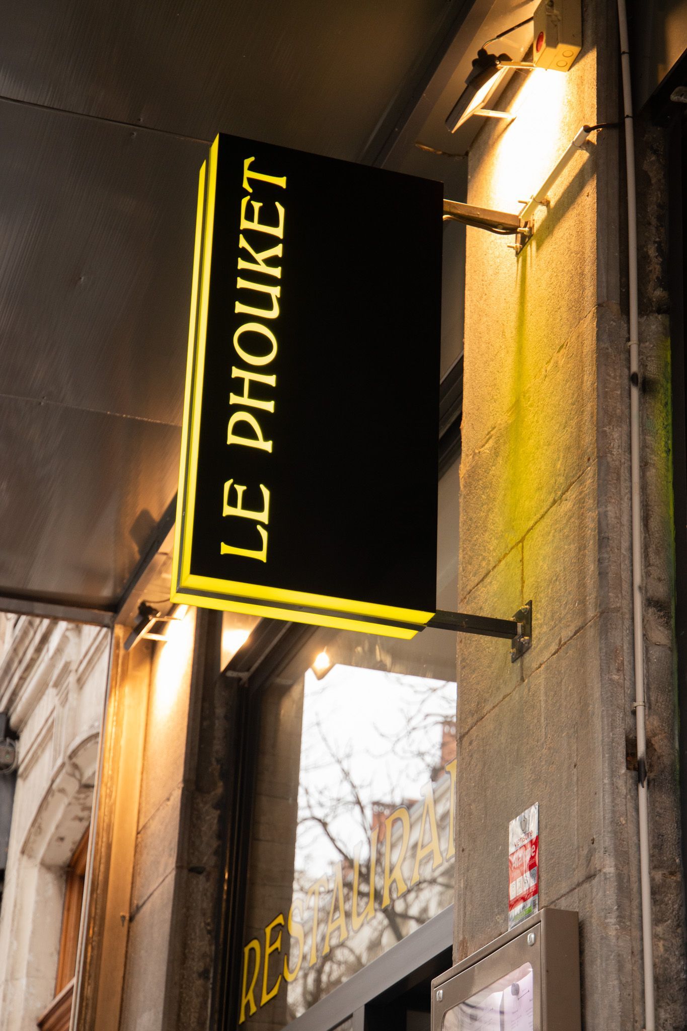 Enseigne rectangulaire noire « LE PHOUKET » avec bordure en néon jaune, fixée sur un bâtiment en béton.