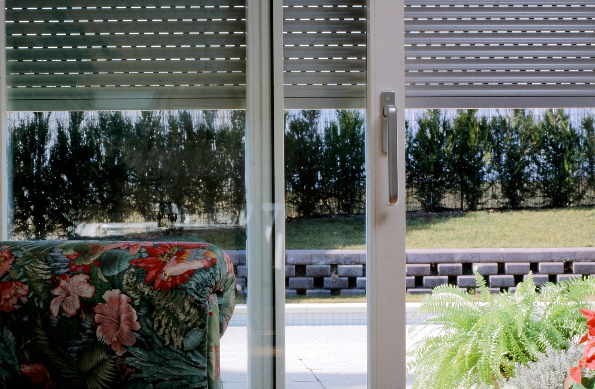 Porte-fenêtre coulissante avec stores fermés, fauteuil à motifs et terrasse ensoleillée avec de la verdure à l'extérieur.