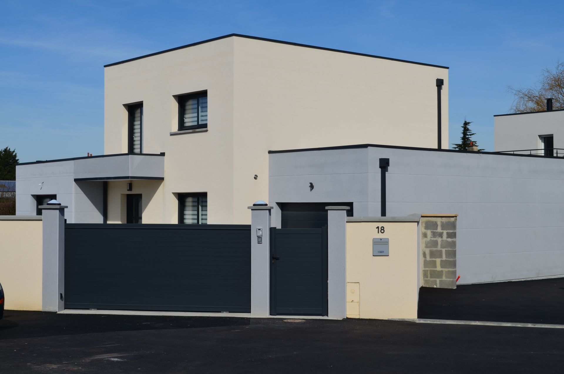 Maison moderne à deux étages avec murs blancs, portail sombre et mur en pierre donnant sur une rue pavée.