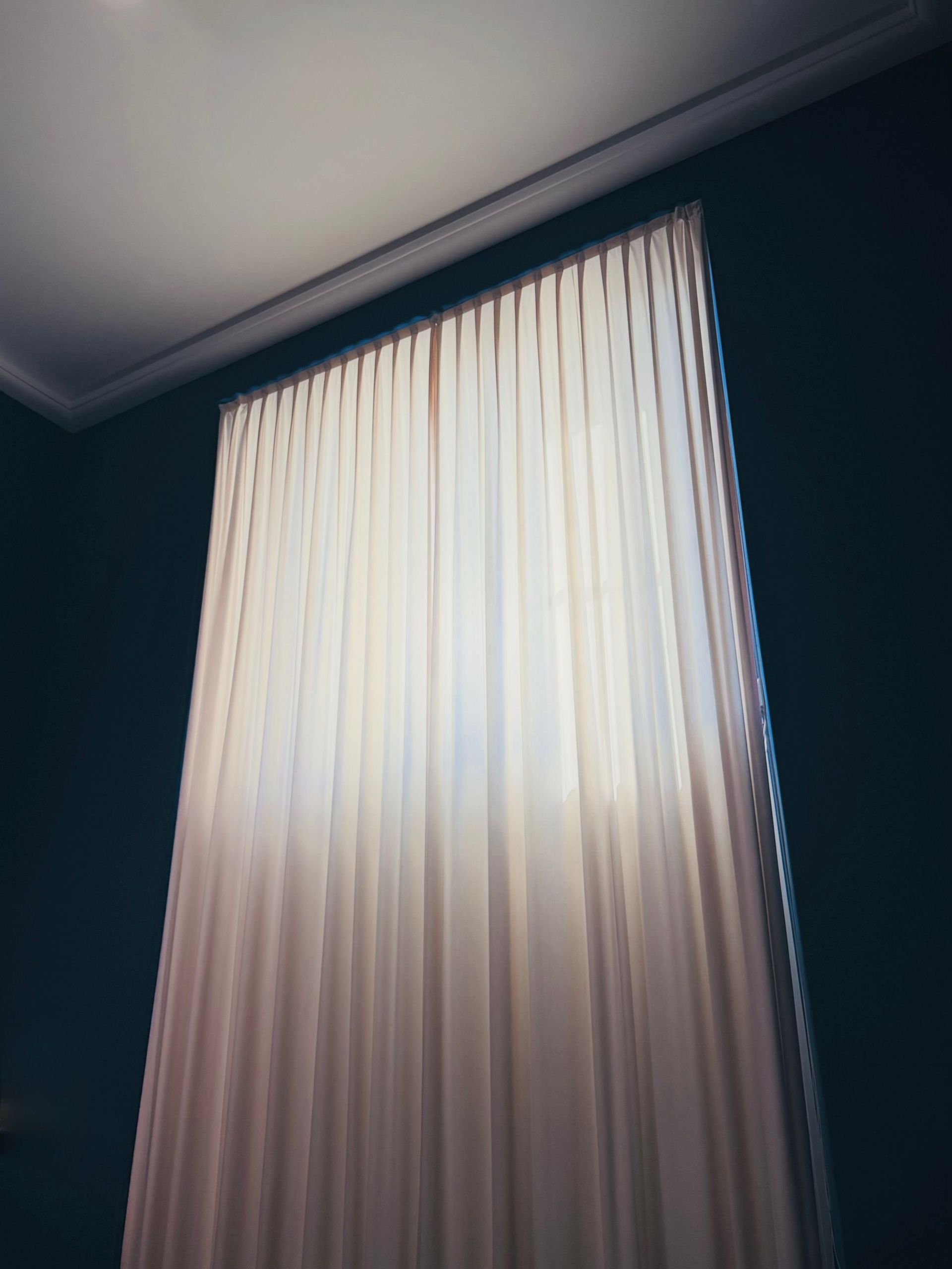 De hauts rideaux crème recouvrent une fenêtre ensoleillée dans une pièce sombre.