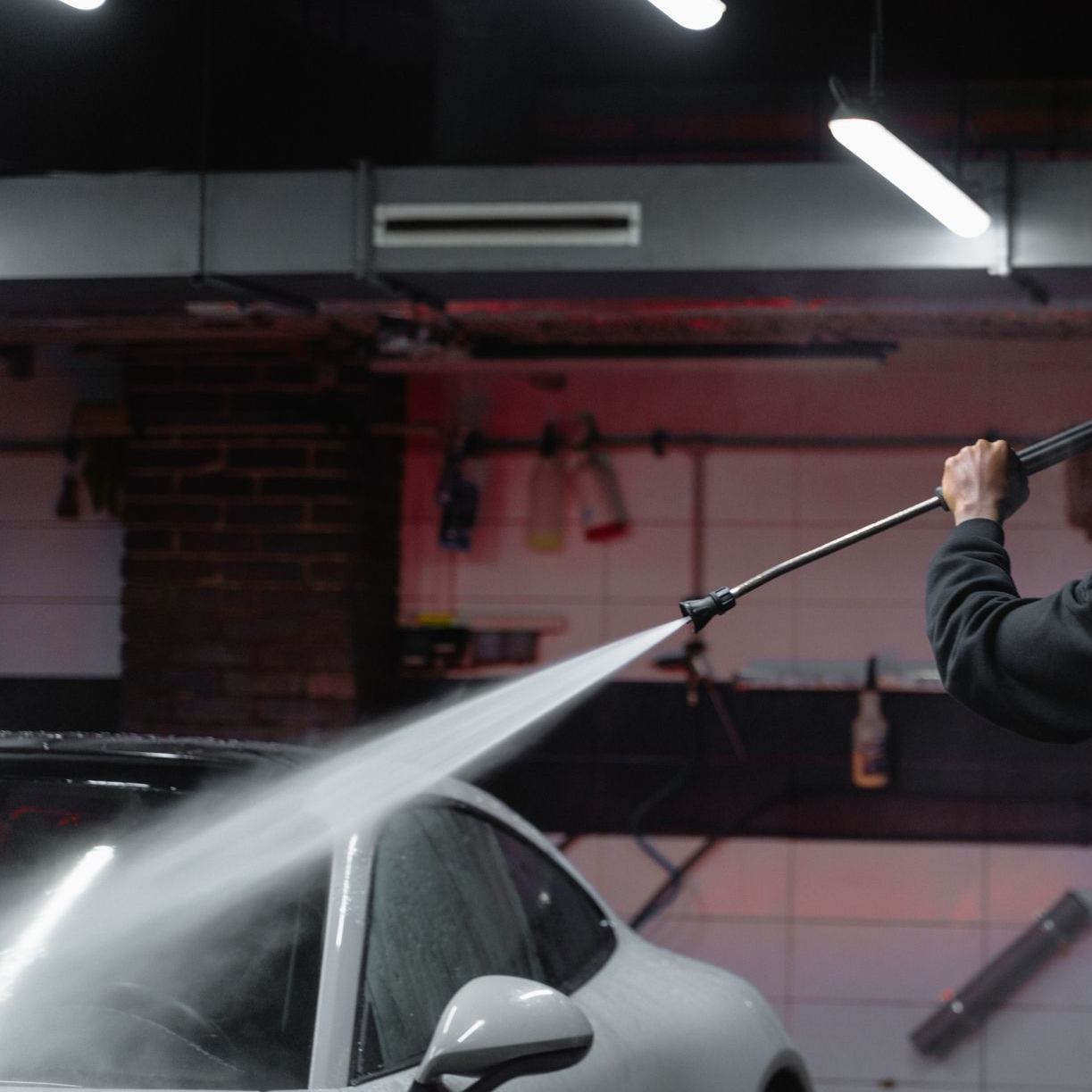 Une personne nettoie une voiture blanche au nettoyeur haute pression à l'intérieur, sous une lumière vive.