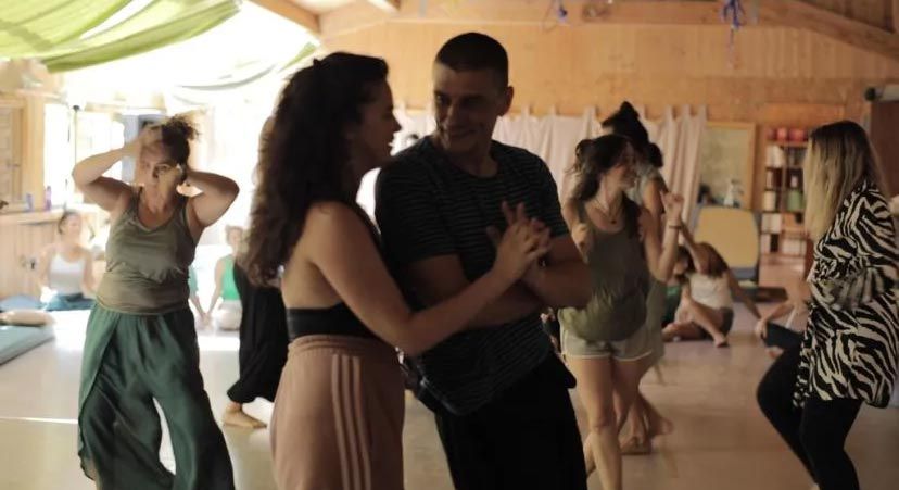 Personas bailando en un cálido estudio de madera, con una pareja en primer plano y otras personas moviéndose detrás