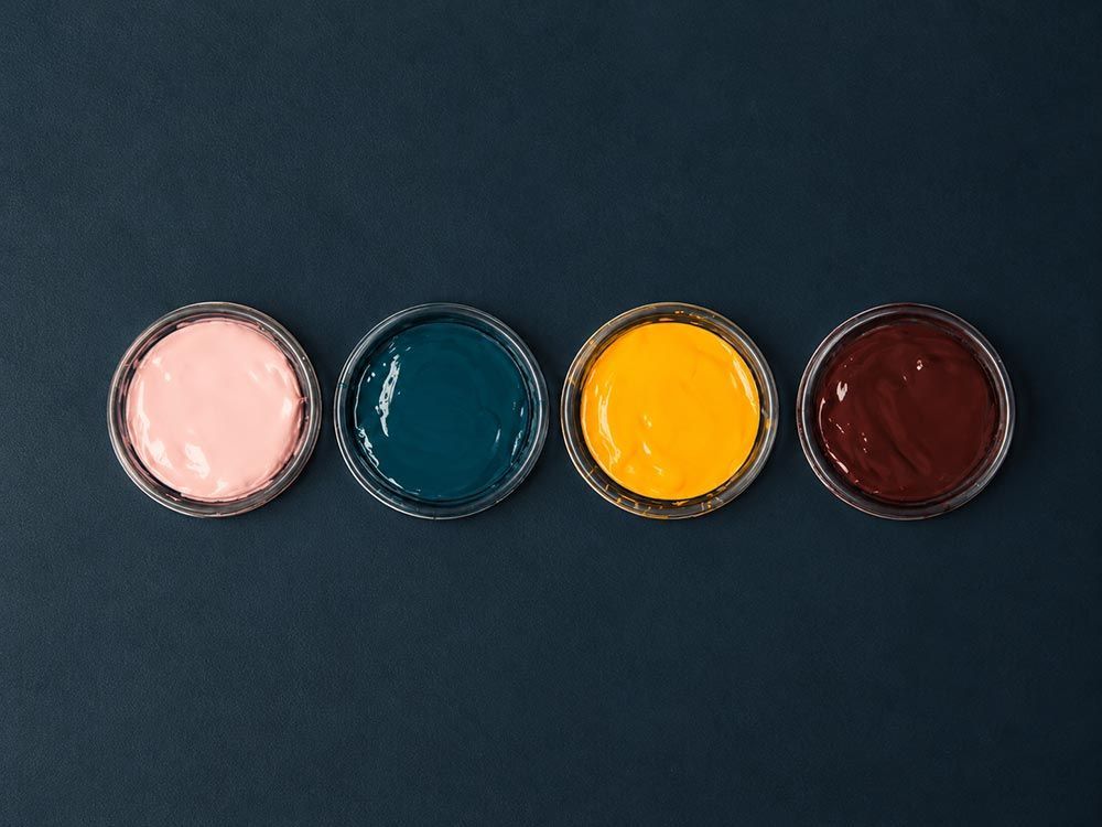 Cuatro pequeños botes de pintura abiertos de color rosa, verde azulado, amarillo y rojo oscuro sobre una superficie azul.