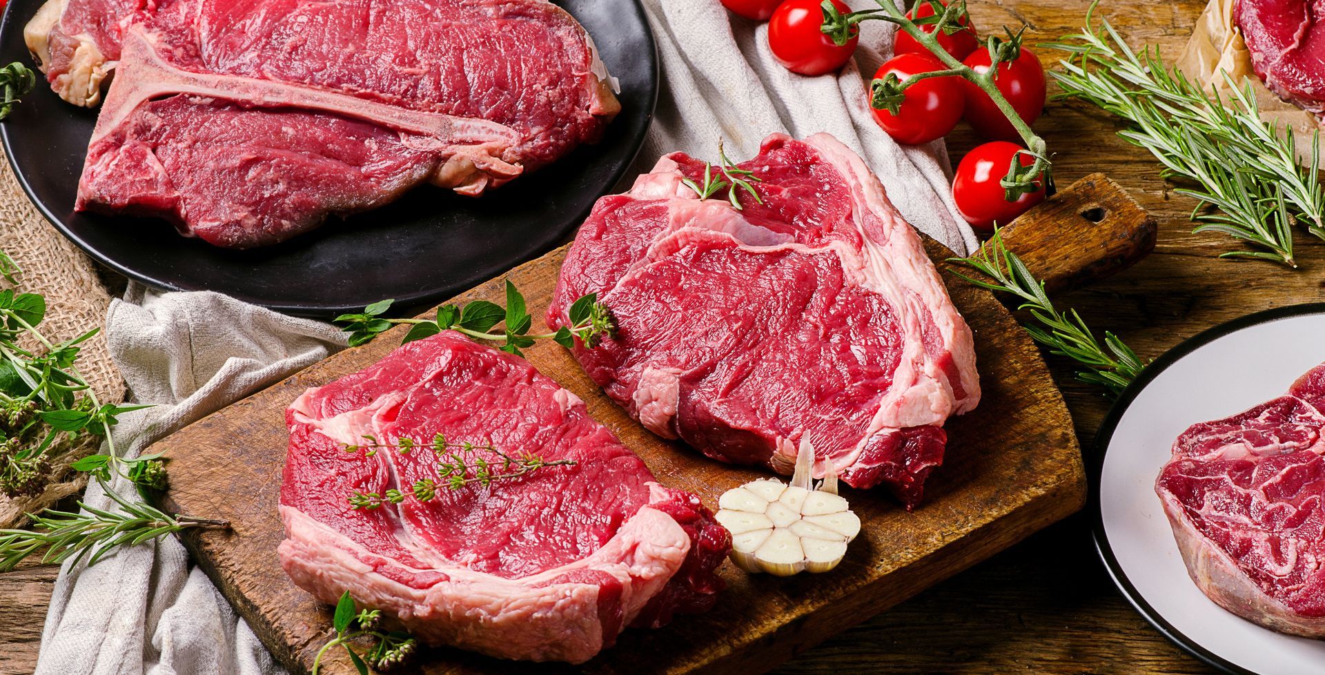 Assortiment de steaks de bœuf crus sur une planche à découper avec de l'ail, des tomates cerises, des herbes et des grains de poivre.