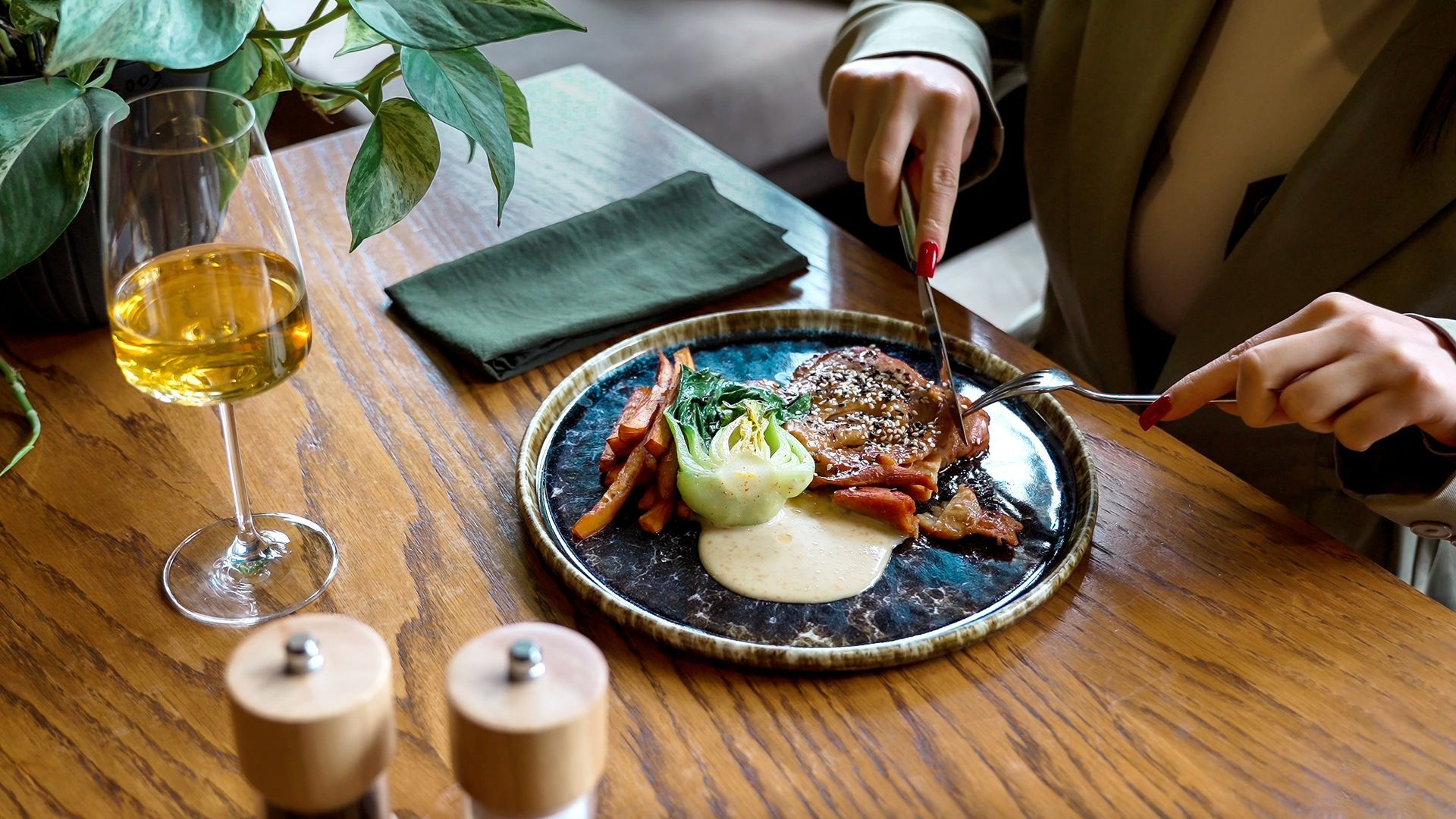 Des mains découpent un steak servi dans une assiette, sur une table de restaurant en bois, accompagné de vin et de condiments.