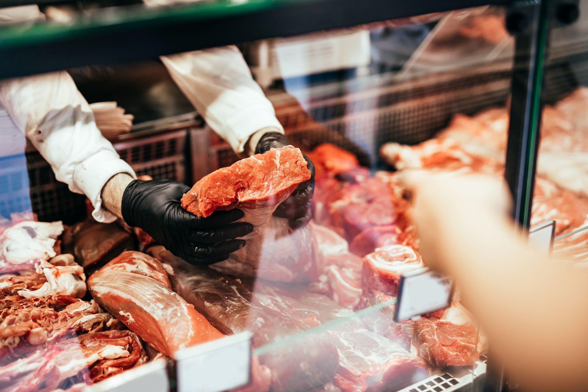 Boucher servant des morceaux de viande crue au comptoir de sa charcuterie, portant des gants noirs et une vitrine réfrigérée.