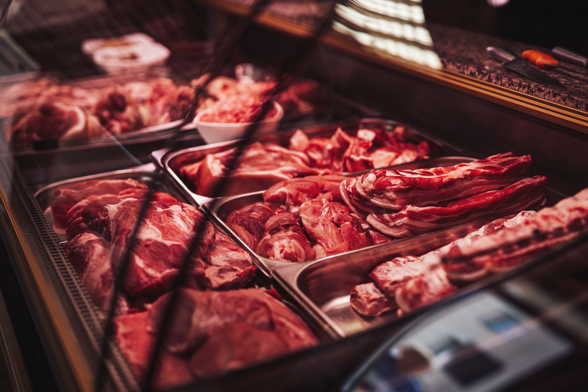 Assortiment de morceaux de viande crue présentés dans une vitrine réfrigérée avec des plateaux et des pinces en métal.