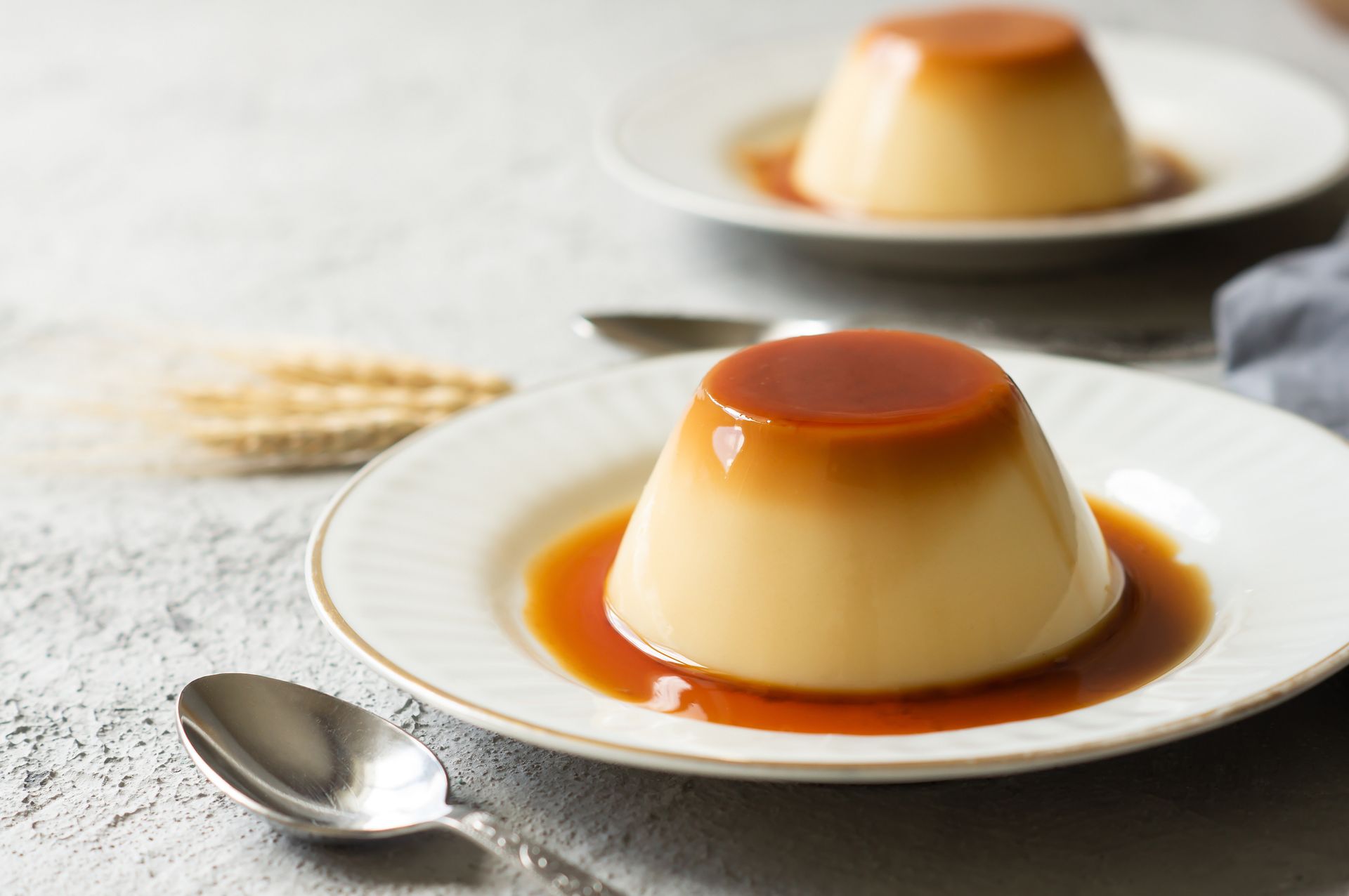 Deux flans au caramel présentés sur des assiettes blanches avec des cuillères, sur une nappe légère.