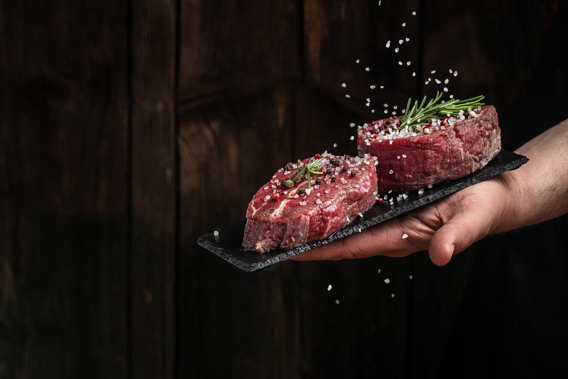 Main tenant deux steaks de bœuf crus sur un plateau noir, garnis d'herbes et de flocons de poivre.