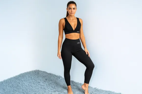 Mujer vestida con un sujetador deportivo negro y mallas, de pie cerca de una pared azul y una alfombra peluda.