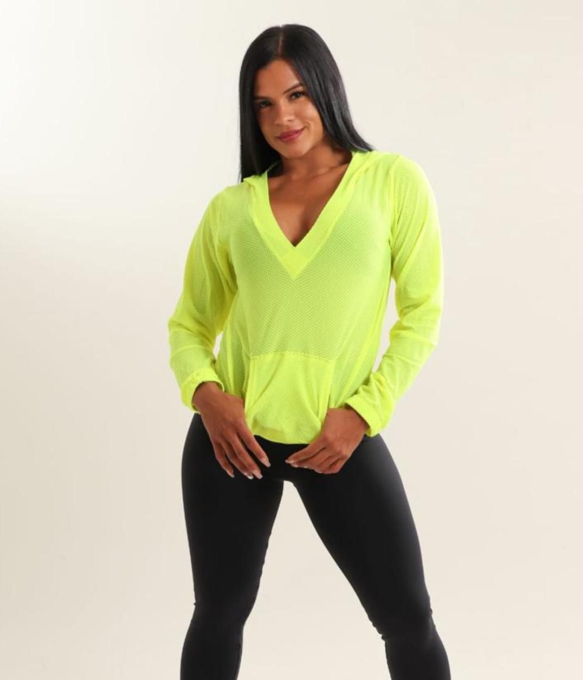 Mujer con capucha verde neón y leggings negros, posando frente a un fondo blanco.