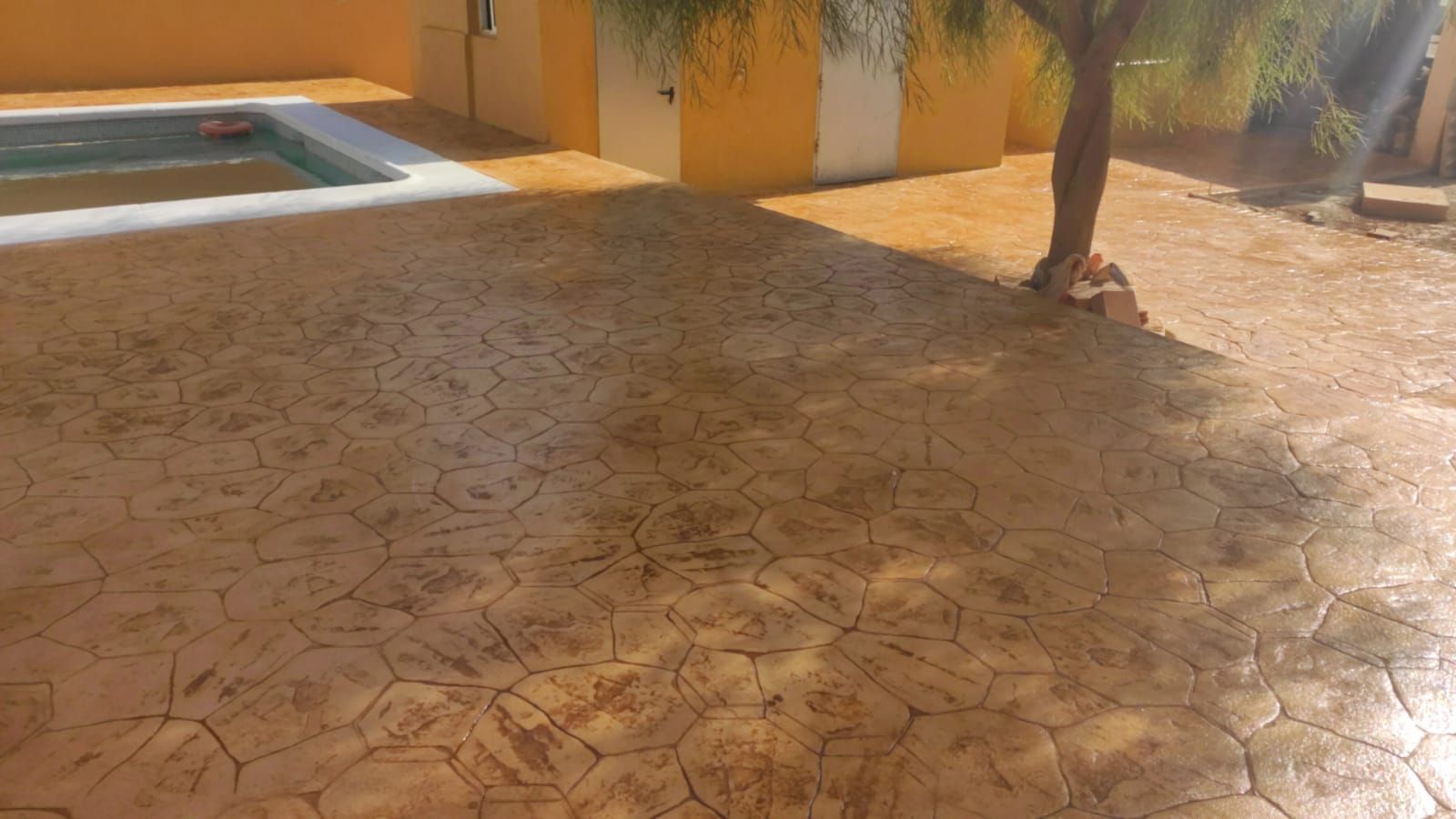 Patio soleado de hormigón estampado con una pequeña piscina, una pared amarilla y un árbol que proyecta sombras.