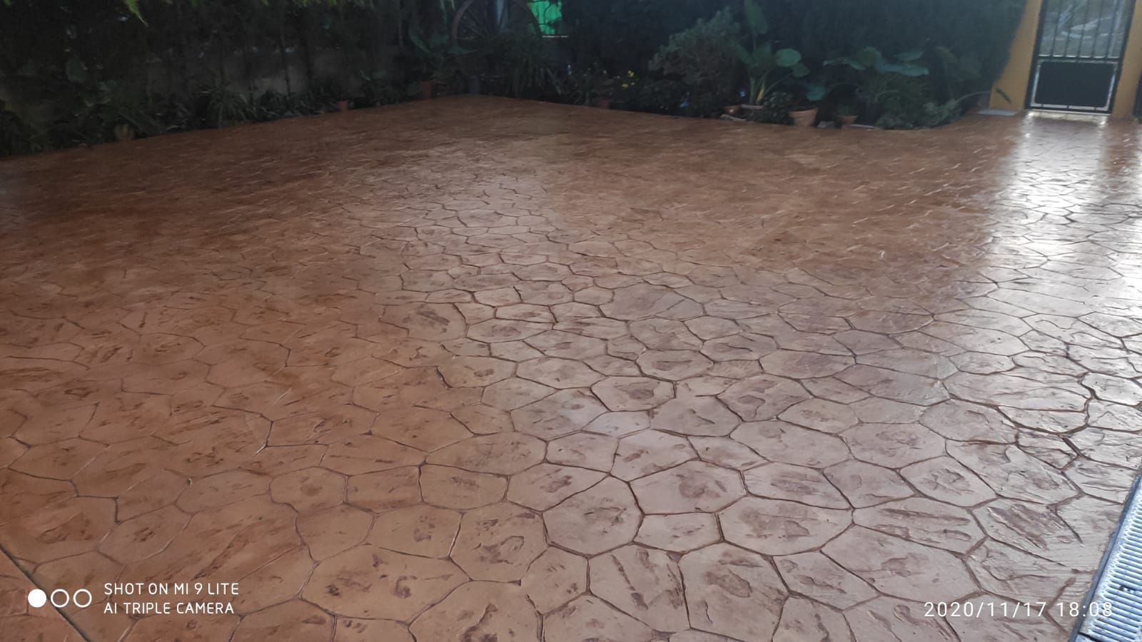 Patio húmedo con baldosas de color marrón rojizo, bajo una zona exterior cubierta, con vegetación al fondo.