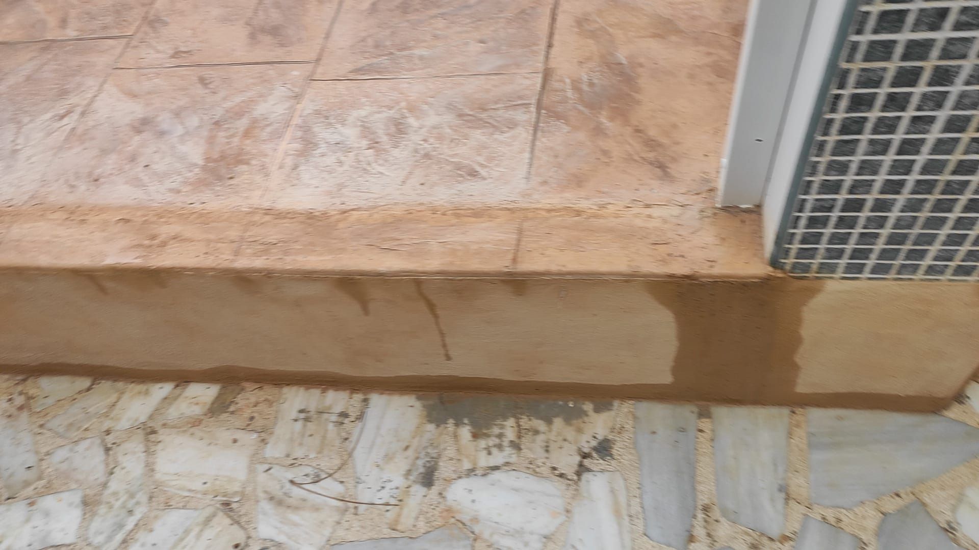 Escalón de baldosas junto a un camino de piedra, con una pared blanca y una rejilla de ventilación metálica a la derecha.