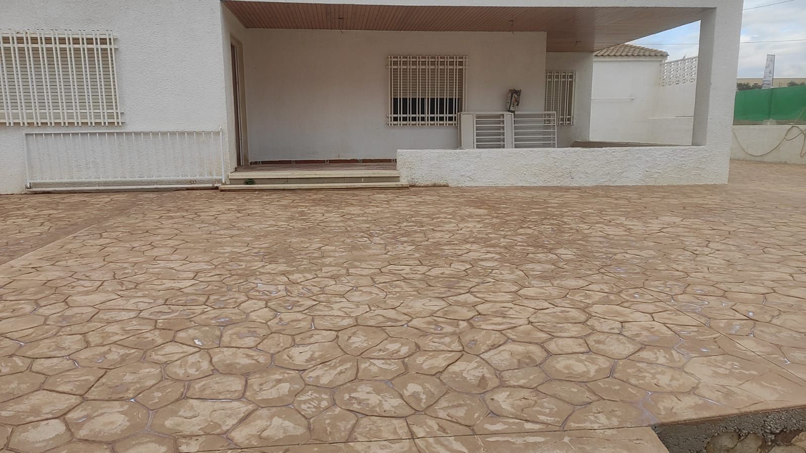 Patio empedrado frente a un edificio blanco con porche cubierto y ventanas enrejadas.