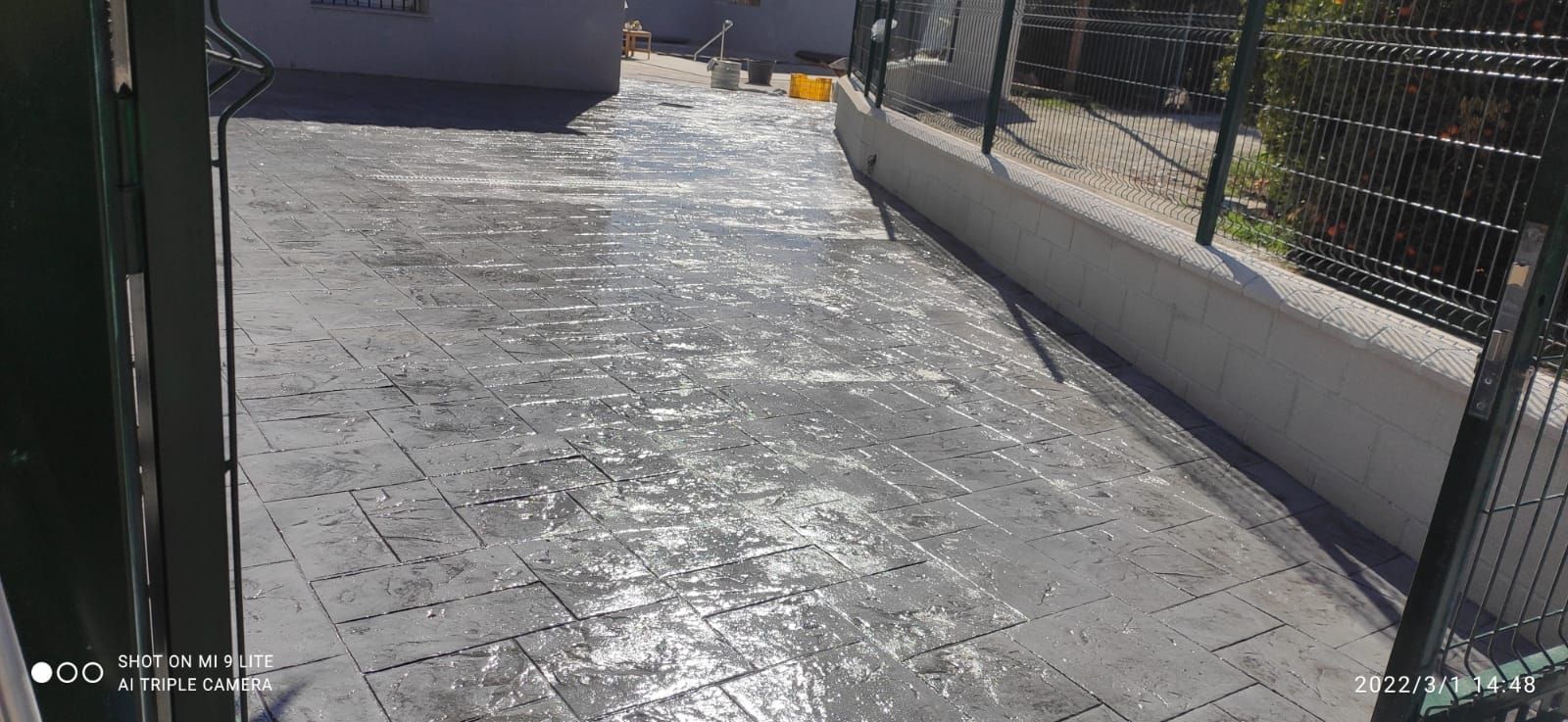 Un soleado camino pavimentado junto a una valla negra y un muro blanco, visto a través de una puerta abierta.