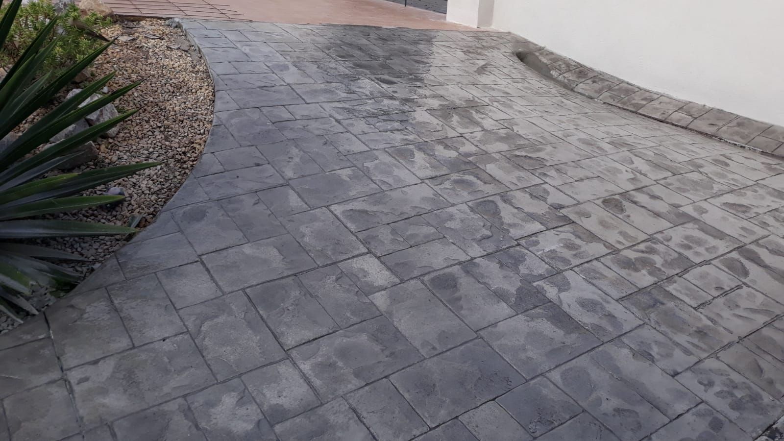 Entrada de vehículos de hormigón estampado gris con pendiente ascendente, junto a un lecho de grava y un muro.