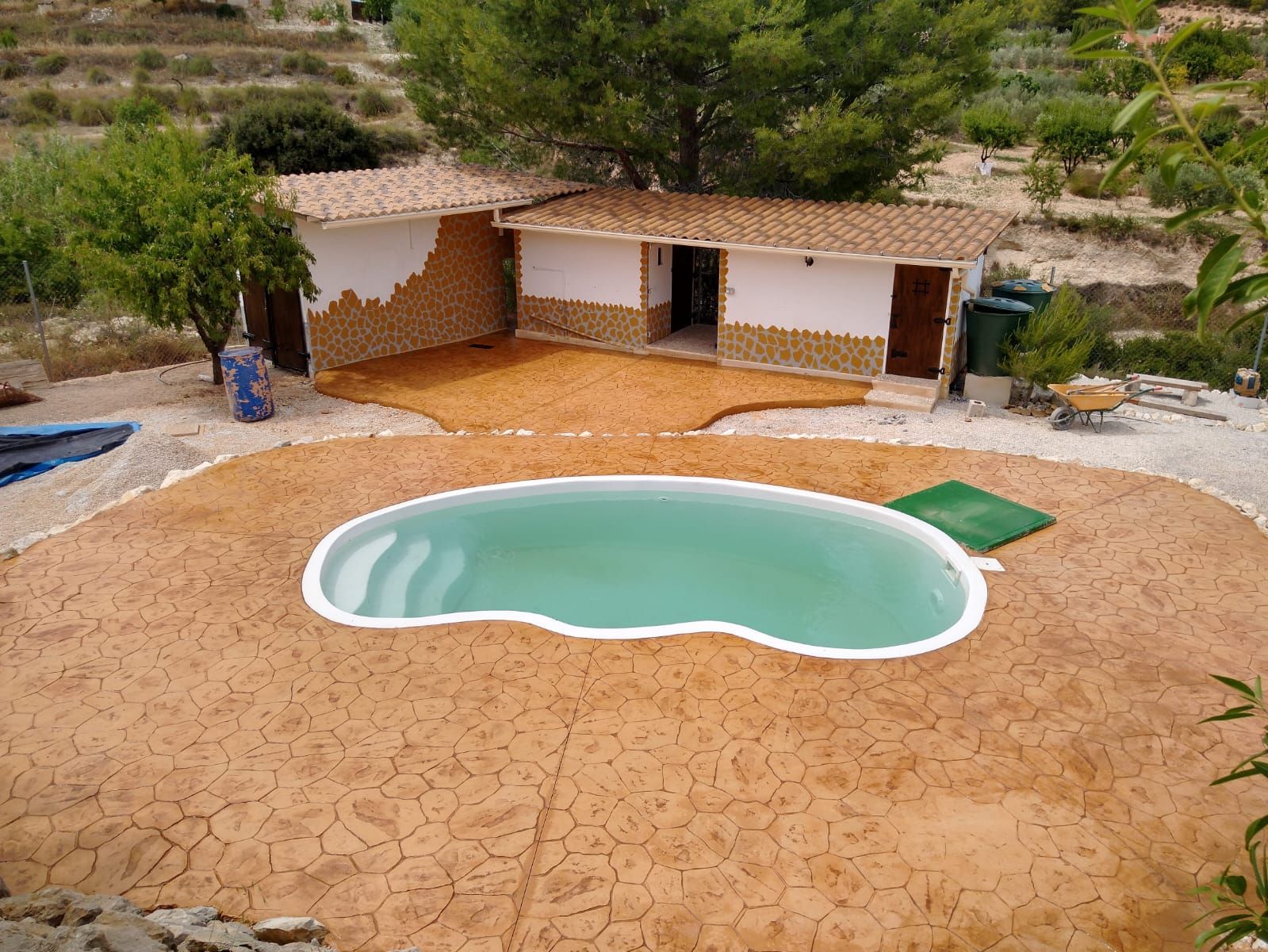 Pequeña piscina en el patio trasero de una casa rústica, rodeada de terrazas de tierra y vegetación.