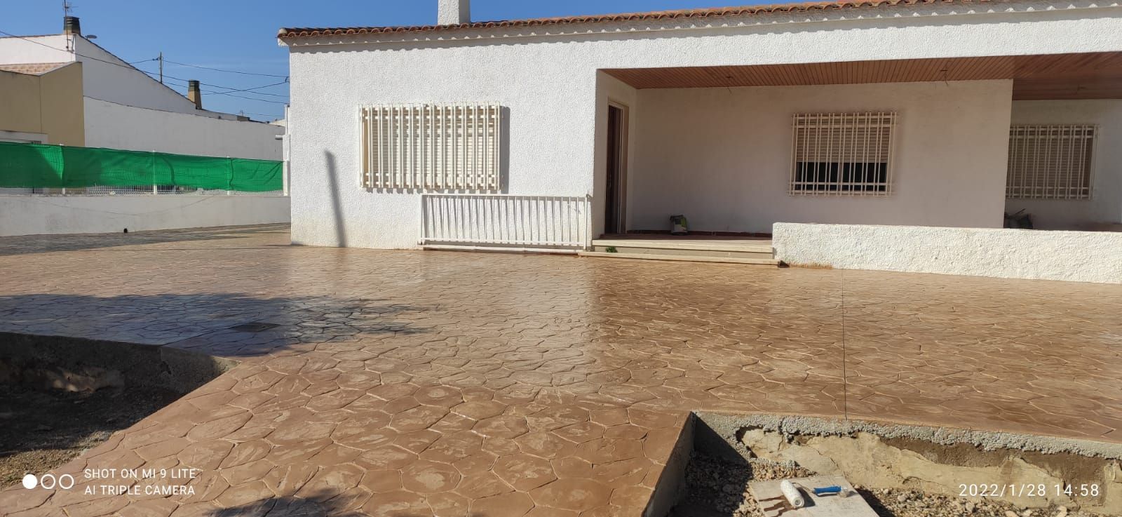 Patio soleado con una casa blanca, ventanas enrejadas y suelo embarrado después de la lluvia.