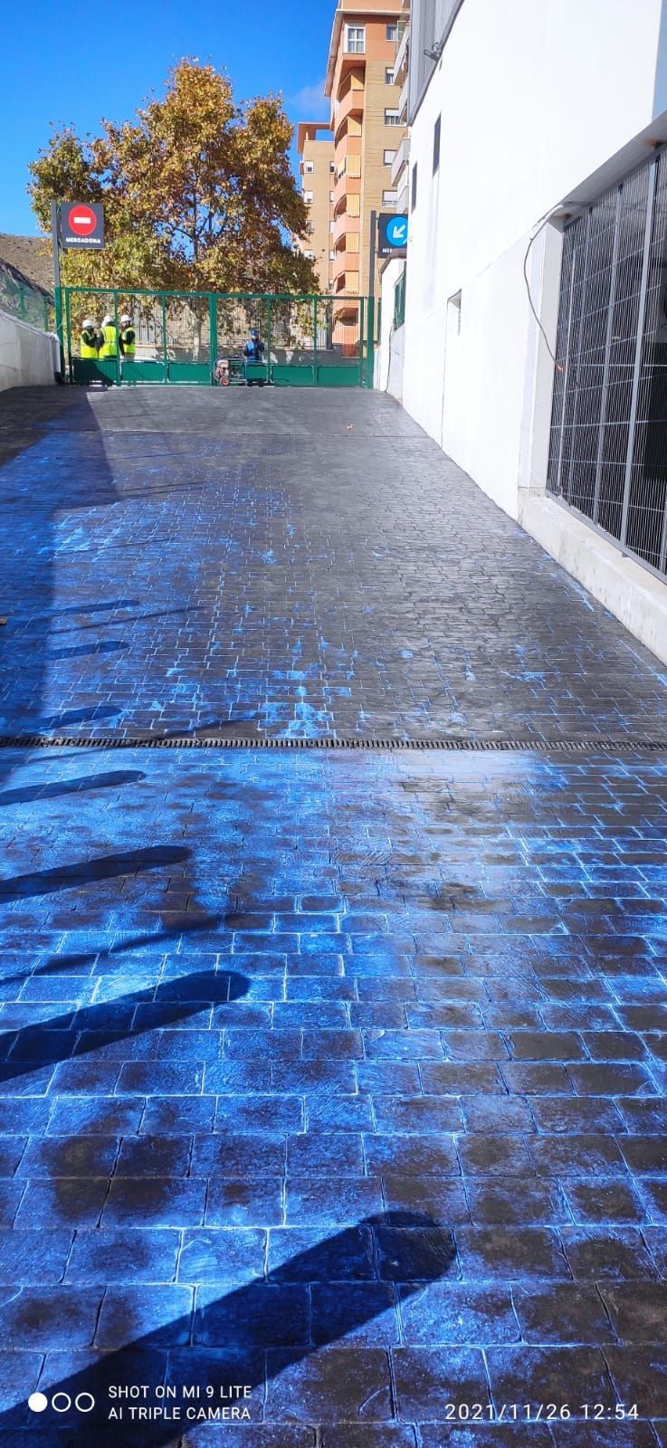 Un sendero mojado de baldosas azules junto a una pared blanca y edificios de apartamentos bajo la luz del sol.