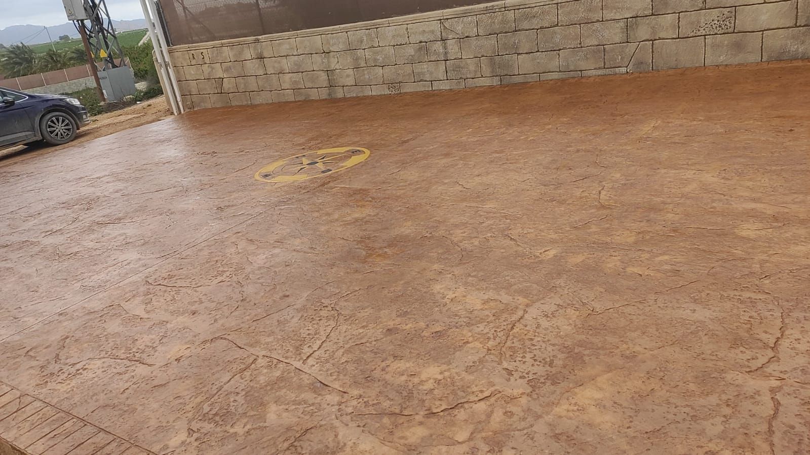 Patio de hormigón vacío con un muro de piedra, suelo texturizado de color marrón anaranjado y un coche aparcado
