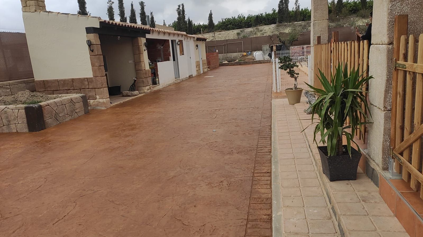 Un estrecho sendero exterior con pavimento rojo, pequeños edificios a la izquierda y plantas en macetas