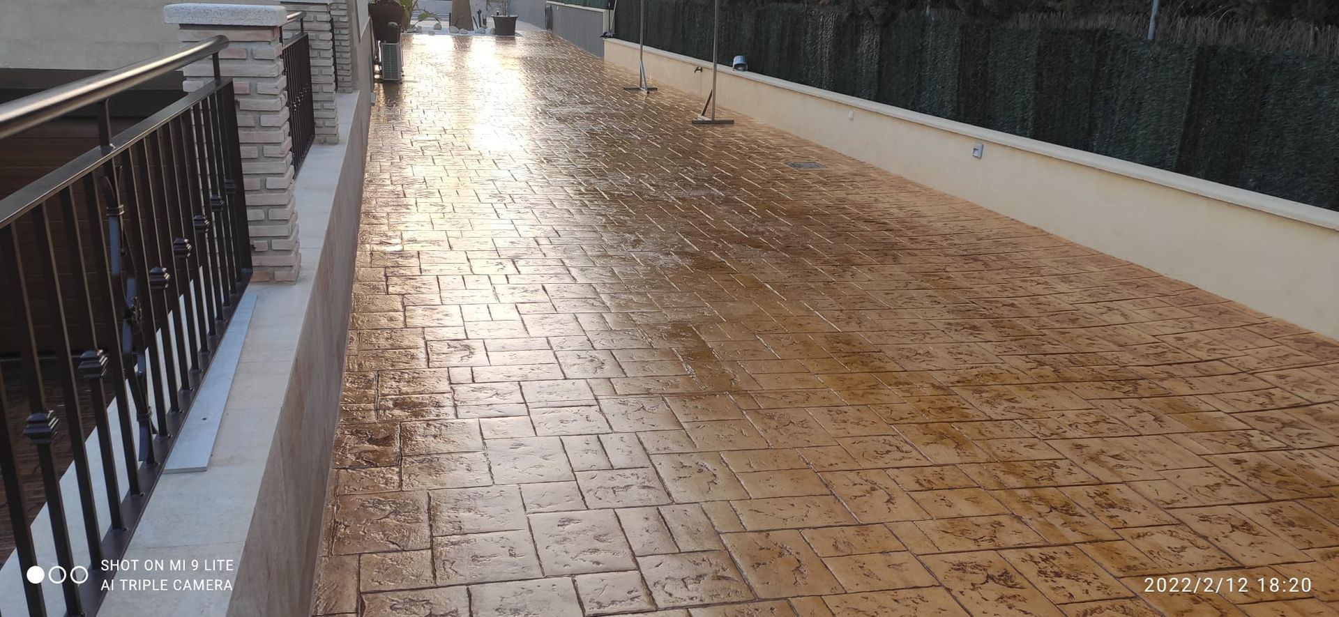 Pasarela de baldosas mojadas junto a una barandilla y un muro, que reflejan la luz después de la lluvia.