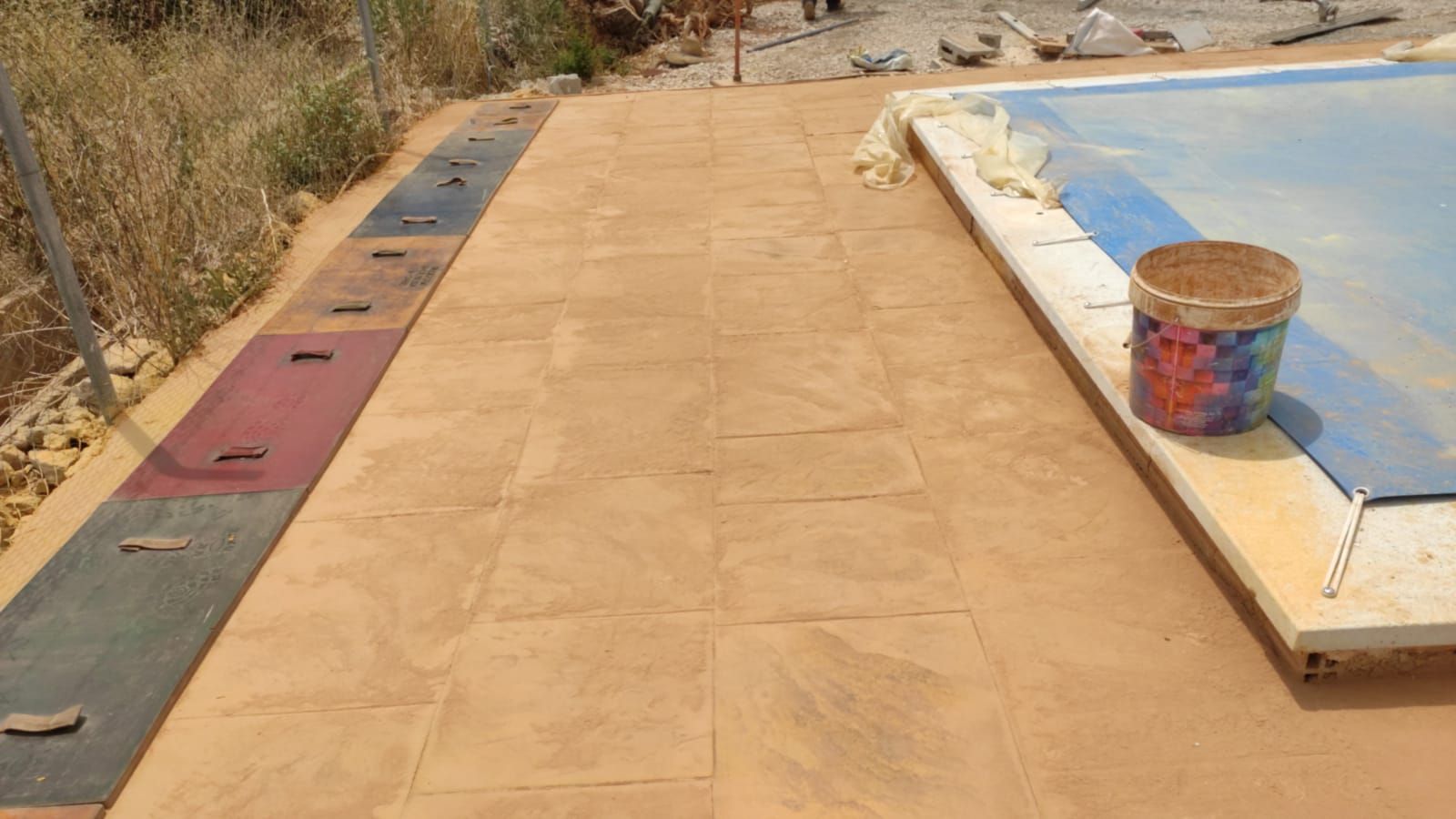Paseo exterior junto a una piscina azul, con un cubo y un bordillo pintado a la derecha.