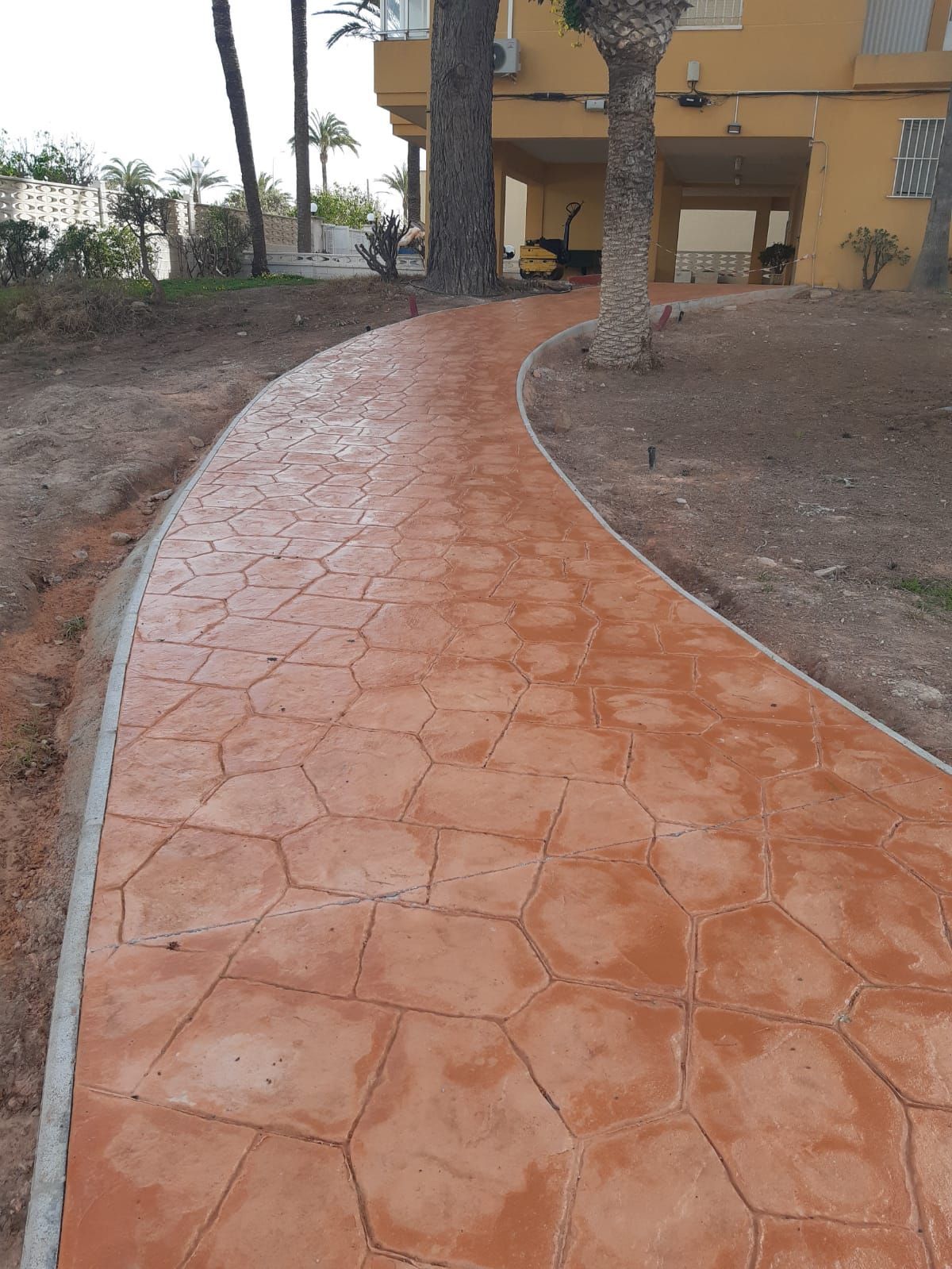 Sendero curvo de terracota que conduce a una casa, bordeado de tierra desnuda y árboles.