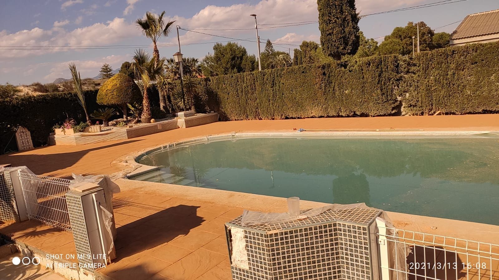 Piscina exterior de agua turquesa, rodeada de un patio de arena, seto y tumbonas bajo un cielo soleado.