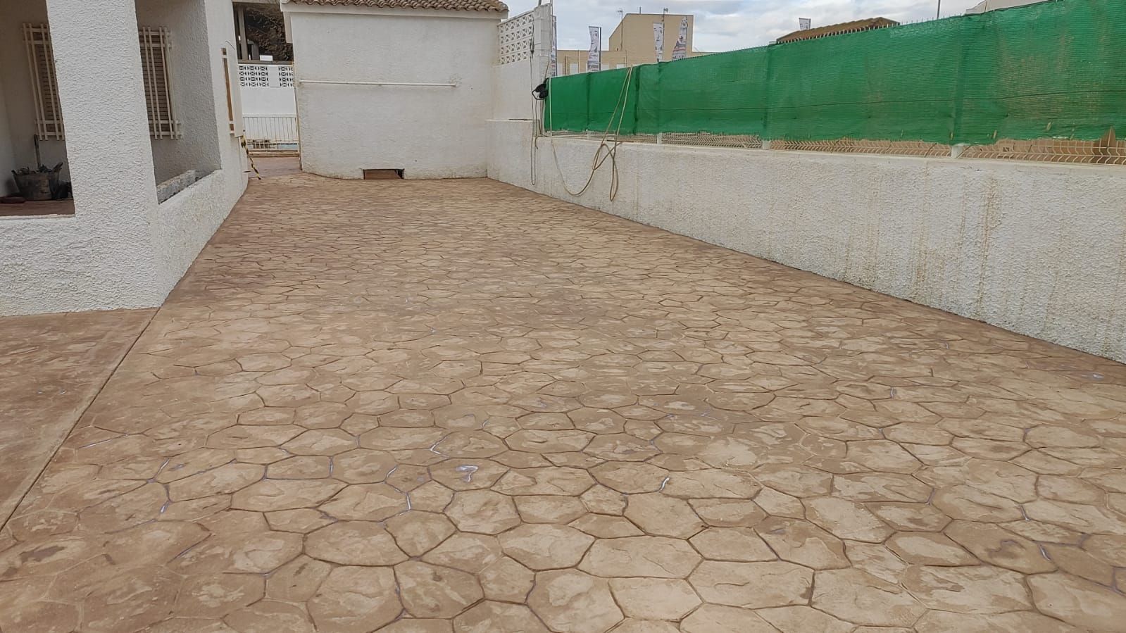 Patio trasero empedrado junto a una casa, delimitado por muros blancos y una valla verde.