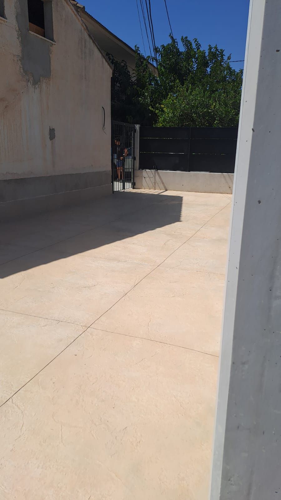 Un soleado paseo al aire libre junto a un edificio, que conduce a una puerta oscura y árboles al fondo.